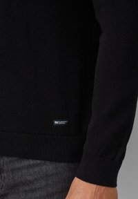 Schwarzer gestrickter Pullover mit langen Ärmeln, gerippten Bündchen und Saum. Verfügt über ein kleines blaues Etikett mit der Aufschrift "Elevated Casual" nahe dem unteren Rand.