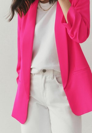 Femme portant un blazer rose vif sur une chemise blanche et un pantalon blanc, avec un collier chaîne en or, posant avec une main près du visage.