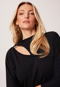 Czarny top z długim rękawem i zakrzywionym dekoltem, z wycięciem i metalicznym akcentem. Gładka faktura i dopasowany krój.