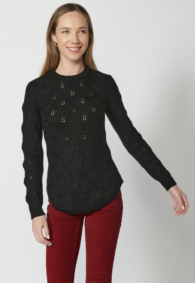 Czarny sweter wykonany z dzianiny z teksturowanym wzorem, długimi rękawami i zaokrąglonym dołem. Noszony z czerwonymi spodniami sztruksowymi.