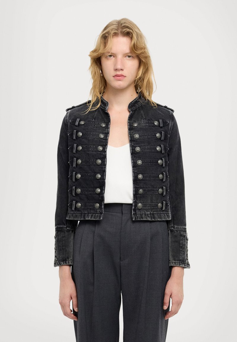 Sort denim cropped jakke med sølvknapper, skulderklapper og høj krave. Båret over en hvid top og parret med grå bukser.