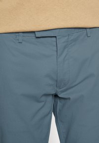 Ljusblå byxor med en slät textur, platt front med en diskret veck och bältesöglor. Tyget verkar lätt och andningsbart.
