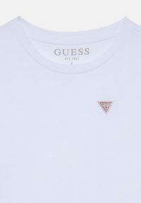 Valge puuvillane lühikeste varrukatega t-särk, millel on vasakul rinnal väike punase ja valge kolmnurga logo ning tekst "GUESS EST. 1981."