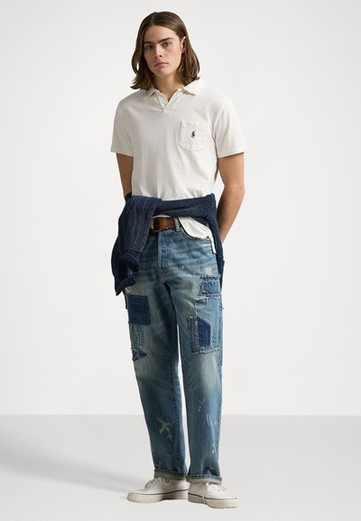 White polo shirt with pocket, light blue distressed jeans with patchwork, and dark sweater tied around waist; casual shoes complete the look.