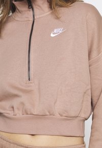 Ljust rosa strapless sweatshirt med hög krage, utrustad med en central dragkedja och broderad Nike-logga på vänster bröst. Mjuk tygtextur.