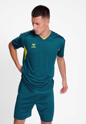 Ung mand, der justerer turkis sportstøj med gule detaljer, står mod en hvid baggrund og kigger nedad.