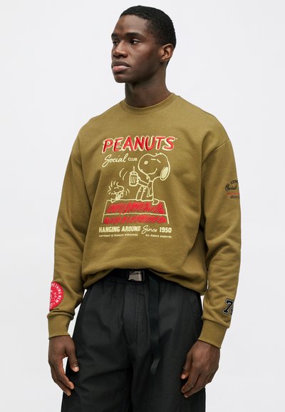 Ελαιοπράσινη μπλούζα με γραφικό "PEANUTS Social Club" και σχέδιο του Σνούπι. Ριμπ άντελφες, χαλαρή εφαρμογή. Μαύρο παντελόνι με κορδόνι.