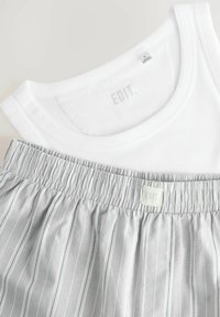 Weißes ärmelloses Oberteil mit geripptem Textur, kombiniert mit hellgrauen Hosen, die vertikale Streifen und einen elastischen Bund haben, beschriftet mit "EDIT."