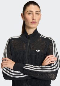 Crna mrežasta jakna na patent s visokim ovratnikom, bijele pruge na rukavima i izvezenim logotipom na prsima.