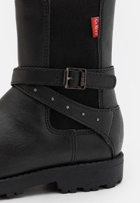 Botín negro de cuero con panel de tejido, detalle de hebilla y tachuelas decorativas. Presenta una suela de goma con un patrón de banda de rodadura resistente.