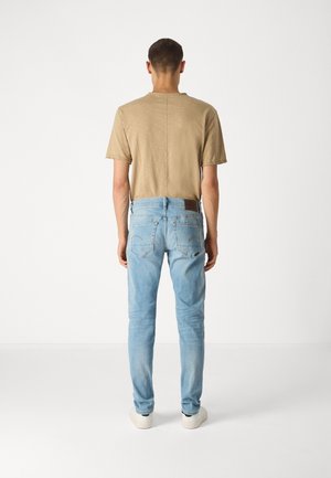 Man die met de rug naar de camera staat, draagt een beige kort shirt, lichtblauwe spijkerbroek en witte sneakers tegen een effen achtergrond.