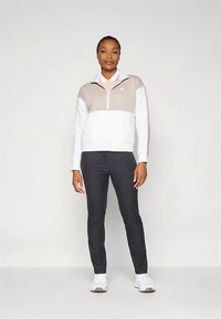 Πουλόβερ με φερμουάρ μέχρι τη μέση σε μπεζ και λευκό, με σχέδιο σε μονόχρωμη περιοχή· συνδυασμένο με μαύρο παντελόνι και λευκά αθλητικά παπούτσια.