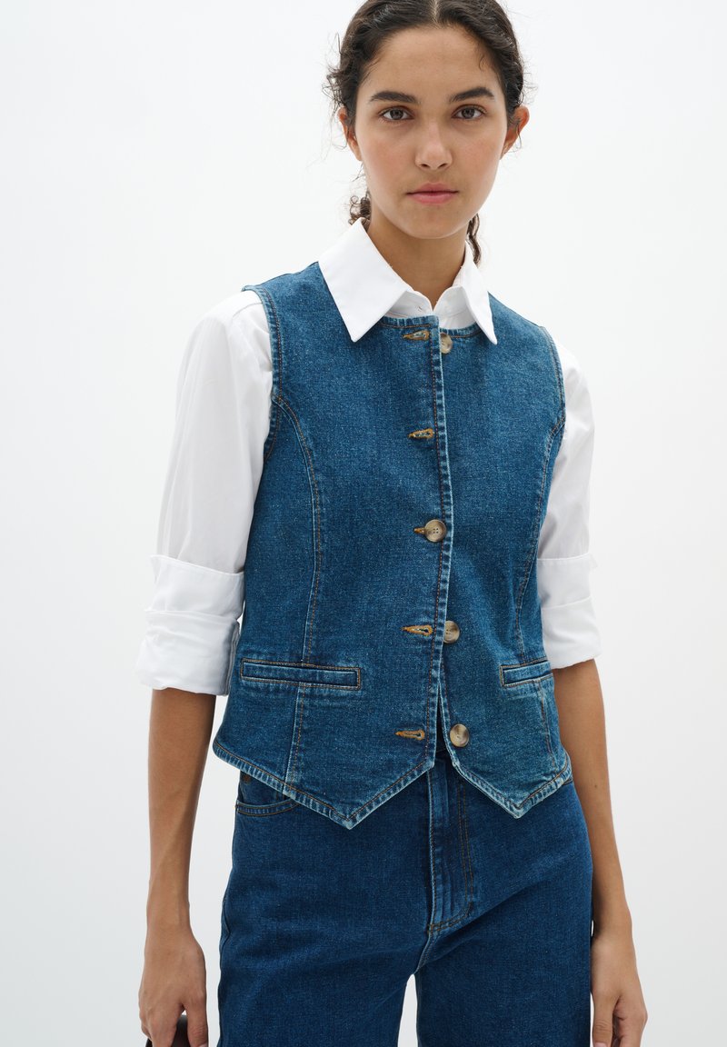 Blauwe denimvest met een knoopsluiting aan de voorkant, twee zijzakken en een aansluitend ontwerp. Gekleed over een witte blouse met kraag.