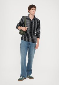 Tricou polo cu maneci lungi, în dungi alb-negru, cu fermoar la gât, asociat cu blugi evazați de un albastru deschis și adidași verzi. Ține o geantă de culoare verde măslin.