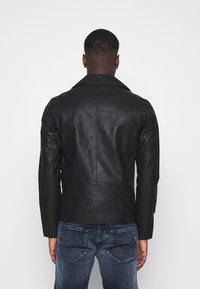 Giacca di pelle nera con trama liscia, colletto pieghevole e design aderente. Caratteristiche includono dettagli cuciti e maniche lunghe, indossata con jeans blu.