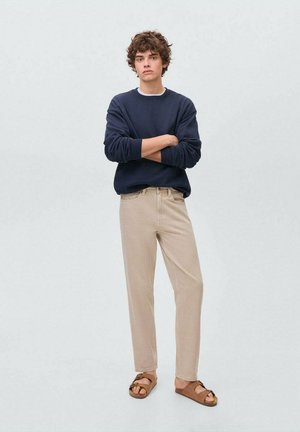 Sweat-shirt marine, pantalon beige et sandales brunes. Le mannequin se tient les bras croisés. La texture douce et la coupe décontractée sont des caractéristiques marquantes.