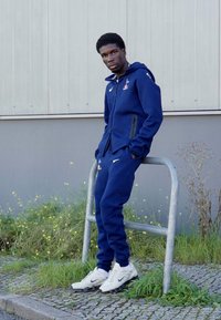 Marineblå træningsdragt i blødt materiale, med en jakke med lynlås og logo samt matchende bukser. Båret med hvide Nike-sneakers.