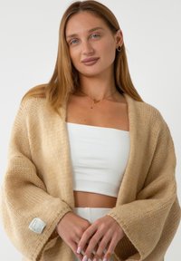 Beżowy kardigan zapinany z przodu z teksturowanym wzorem, szerokimi rękawami i metką na lewym rękawie. Model ma na sobie biały crop top.