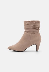 Botín beige informal hasta el tobillo con punta afilada, confeccionado en un material suave similar al ante, con un tacón de altura media y detalles de puño fruncido.