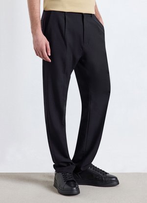 Mann steht, trägt schwarze Hose, schwarze Sneakers und ein beiges Hemd mit Händen in den Taschen vor einem einfarbigen weißen Hintergrund.