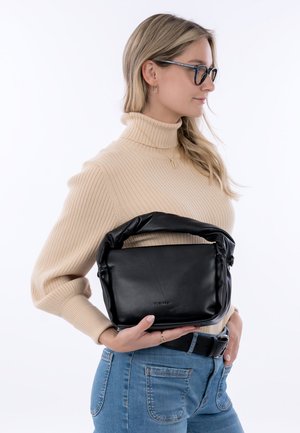 Žena s brýlemi, oblečená v béžovém roláku a modrých džínách, drží černou koženou kabelku s polstrovaným uchy na úrovni pasu.