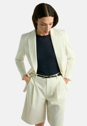 Kobieta ubrana w kremową marynarkę i pasujące plisowane szorty z czarnym paskiem oraz granatową bluzkę, ręce w kieszeniach, patrząca w dół.