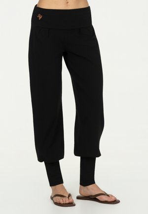 Frau trägt schwarze, geschichtete, weit geschnittene, kurze Hosen mit enganliegenden Leggings darunter und braune Flip-Flops.
