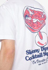 Osoba w białej koszulce z grafiką czerwonego kieliszka koktajlowego, przedstawiającą kobietę pływającą oraz niebieskim napisem "Skinny Dippin' Cocktail Sip."