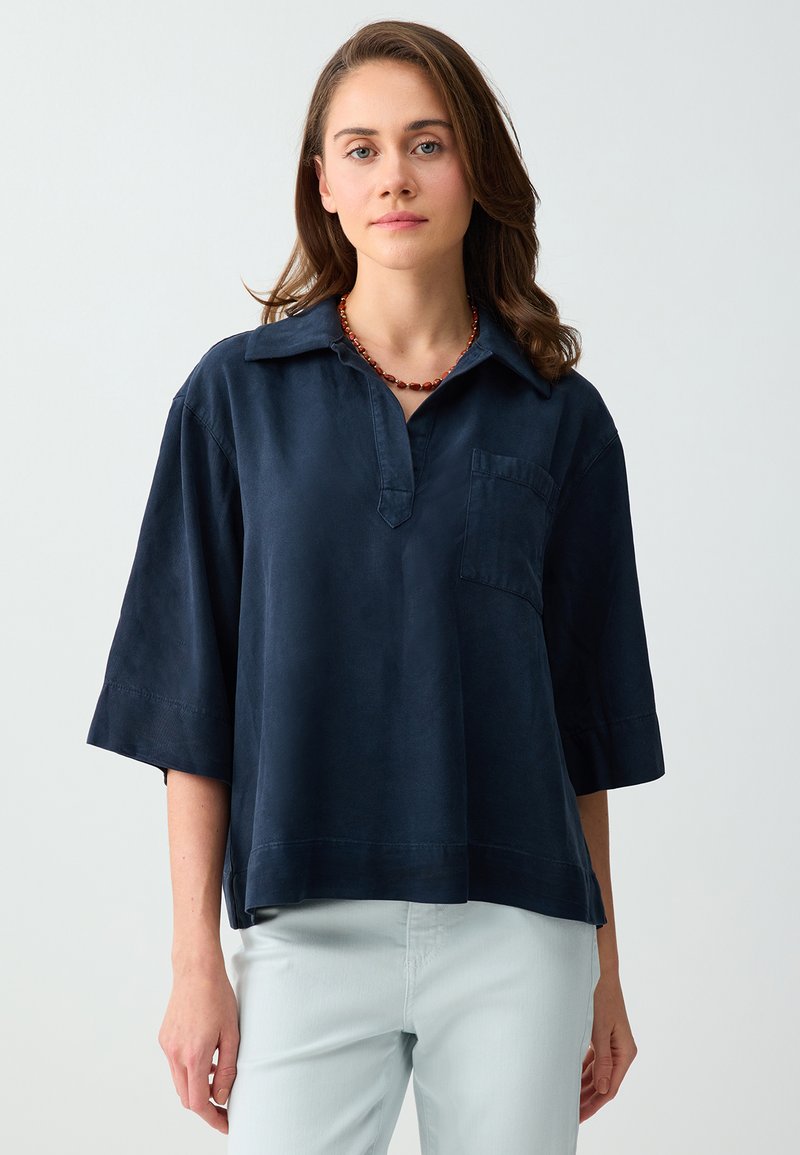 Jeune femme aux cheveux bruns mi-longs, portant une chemise ample bleu marine à col et un pantalon clair sur un fond uni.