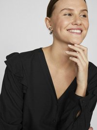 Crna bluzica s volanima na ramenima, V-izrezom i detaljima na dugmadima. Teksturalna tkanina s glatkom završnom obradom. Model se dodiruje po bradi.