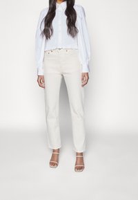 Blusa blanca texturizada con mangas largas abullonadas y dobladillo en forma de scallop, combinada con pantalones rectos blanco roto y sandalias con tiras transparentes.