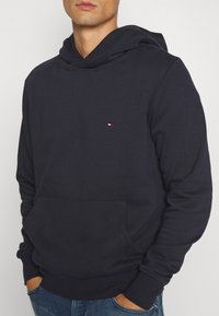 Tmavě modrá bavlněná mikina s kapucí, přední kapsou ve stylu klokanky, žebrovanými manžetami a lemem, s malým vyšitým logem na hrudi.