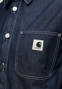 Σκούρο μπλε πουκάμισο τζιν με μπροστινή τσέπη, λευκά γαζιά και τετράγωνη ετικέτα με το λογότυπο Carhartt στην τσέπη. Διαθέτει μεταλλικά κουμπιά.