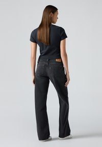 Mujer con cabello largo castaño, vistiendo una camiseta negra de manga corta y jeans negros de cintura alta, de pie contra un fondo claro liso, de espaldas.