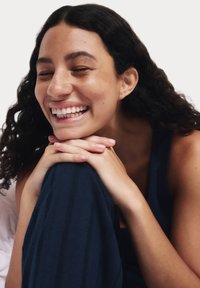 Persona sonriente con cabello negro rizado, apoyando el mentón en las manos y vistiendo una camiseta azul marino. La piel es clara y está salpicada de pecas.