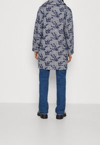 Persoon in een blauw-wit gestreepte jas, met blauwe spijkerbroek en zwarte schoenen, staande tegen een effen witte achtergrond, met de rug naar de camera gekeerd.