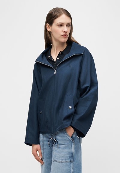 Γυναίκα που φοράει ναυτικό μπλε μπουφάν με φερμουάρ πάνω από μαύρο πουκάμισο με γιακά και ανοιχτό μπλε τζιν, στέκεται με το δεξί της χέρι στην τσέπη.