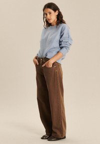 Jasnoniebieski sweter robiony na drutach z prążkowanym wykończeniem, zestawiony z brązowymi spodniami o wysokiej talii i szerokich nogawkach. Model nosi ciemne buty i ma ręce w kieszeniach.