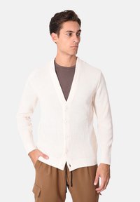 Cardigan crema, tessuto a coste, scollo a V profondo, cinque bottoni, spaccature laterali, indossato sopra una t-shirt marrone. Vestibilità casual con maniche lunghe.