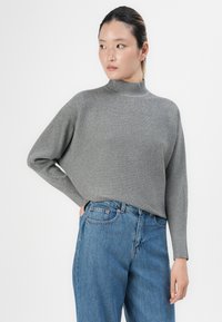 Woman wearing a gray ribbed turtleneck sweater and blue high-waisted jeans, standing with one hand in pocket and looking sideways.