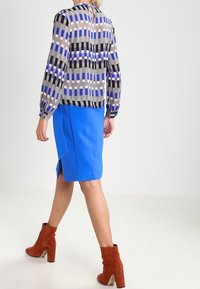 Kvinna som går bort med en mönstrad långärmad blus, ljusblå knälång kjol med sprund, och rödbruna klackade ankelstövlar.