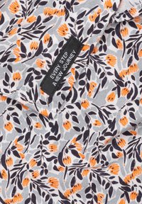 Bloemenpatroon stof in grijs met oranje bloemen en donkere bladeren. Een zwart etiket met de tekst "IEDERE STAP EEN NIEUWE REIS." Zachte textuur van de stof.