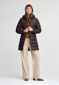 Abrigo puffer marrón con capucha, que cuenta con cierre de cremallera, acolchado horizontal y un logotipo bordado. Combinado con pantalones de pierna ancha beige.