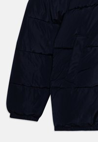 Mouw van een marineblauwe donsjas, met een doorgestikt ontwerp, elastische boorden en een zijzak. Het materiaal lijkt glad en synthetisch.