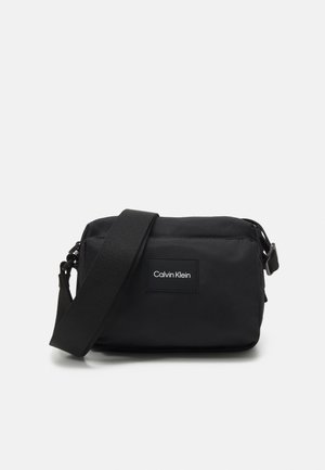 Schwarze kompakte Crossbody-Tasche mit breitem verstellbarem Riemen und Calvin Klein Logo-Patch vorne.