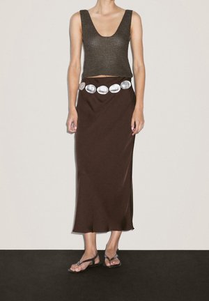 Kobieta ubrana w bezrękawnikowy krótki sweter, ciemnobrązową spódnicę midi z srebrnym owalnym paskiem oraz płaskie sandały na paski, stojąca na tle gładkiej ściany.