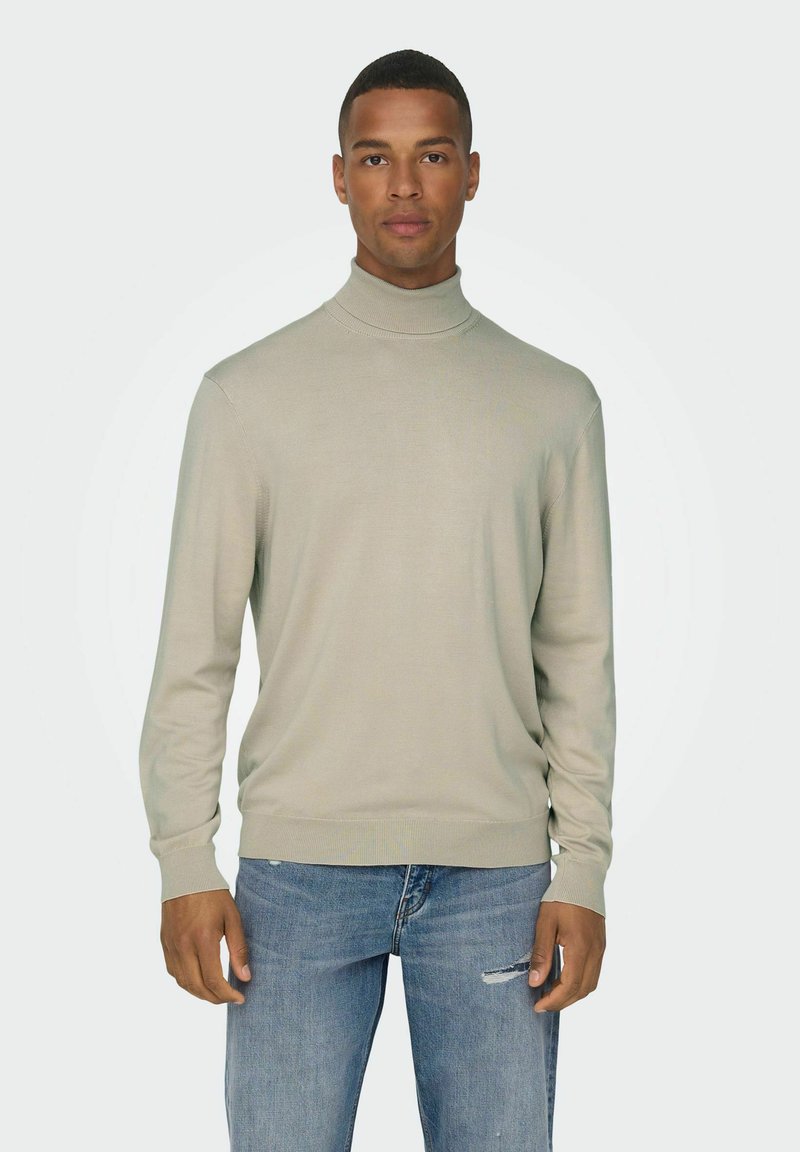 Svetlo siva turtleneck pulover iz mehkega teksturiranega materiala, z dolgimi rokavi in prilegnjenim dizajnom. Usklajen z modrimi uničenimi kavbojkami.