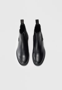 Une paire de bottes Chelsea en cuir noir avec des panneaux élastiques sur les côtés et des tirants, sur un fond blanc, vues du dessus.