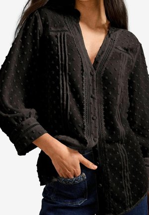 Femme portant un chemisier noir texturé à boutons avec des détails en dentelle et des manches bouffantes, la main dans la poche avant d'un jean bleu foncé.