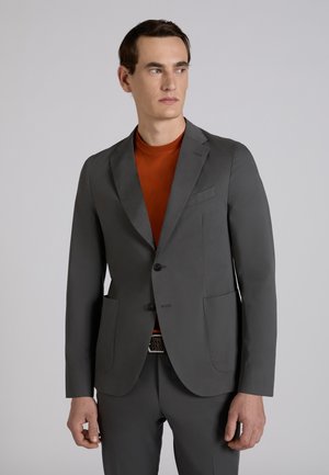 Man in donkergrijs pak en een oranjebruine overhemd staand tegen een lichtgrijze achtergrond, kijkend naar rechts.